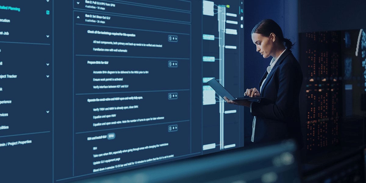 Engineer testing digital well operations software on a laptop with workflow dashboards and analytics on large screens in a control room.